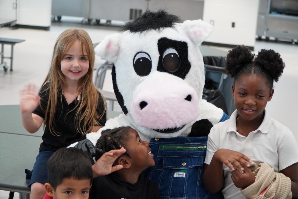 A photo of a cow mascot with a group of kids around him.