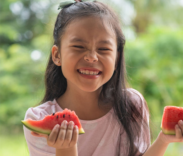 An Asian American child smiling big, while holding a watermelon.