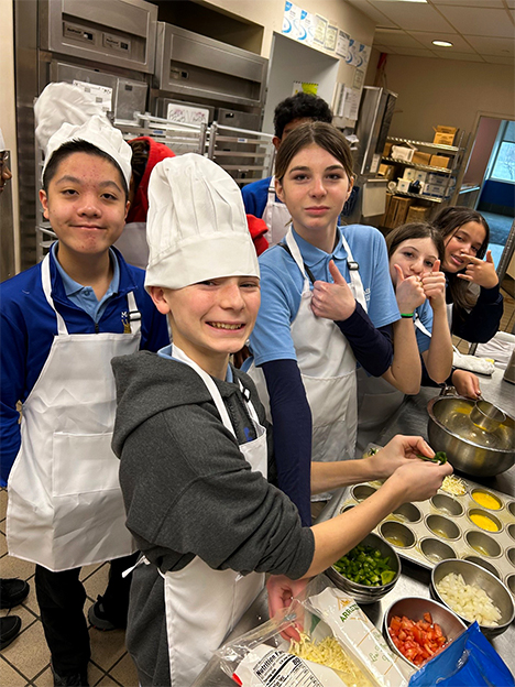 A group of kids in the kitchen wearing aprons.