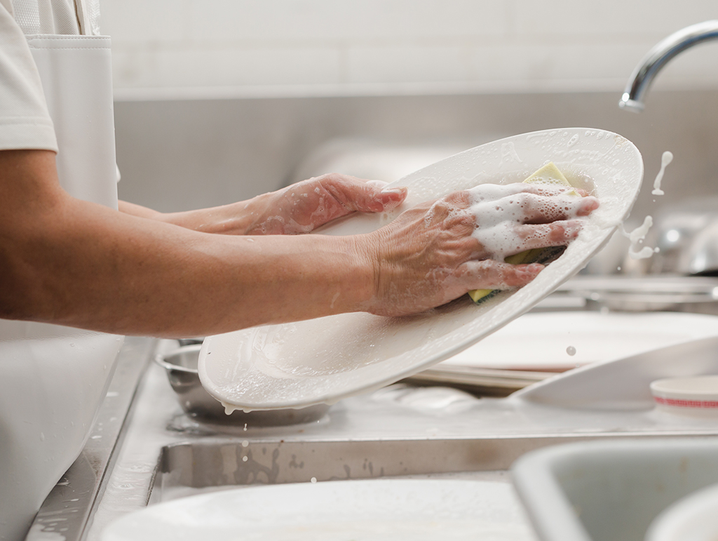 A person over a sink hand washing a white dinner plate.