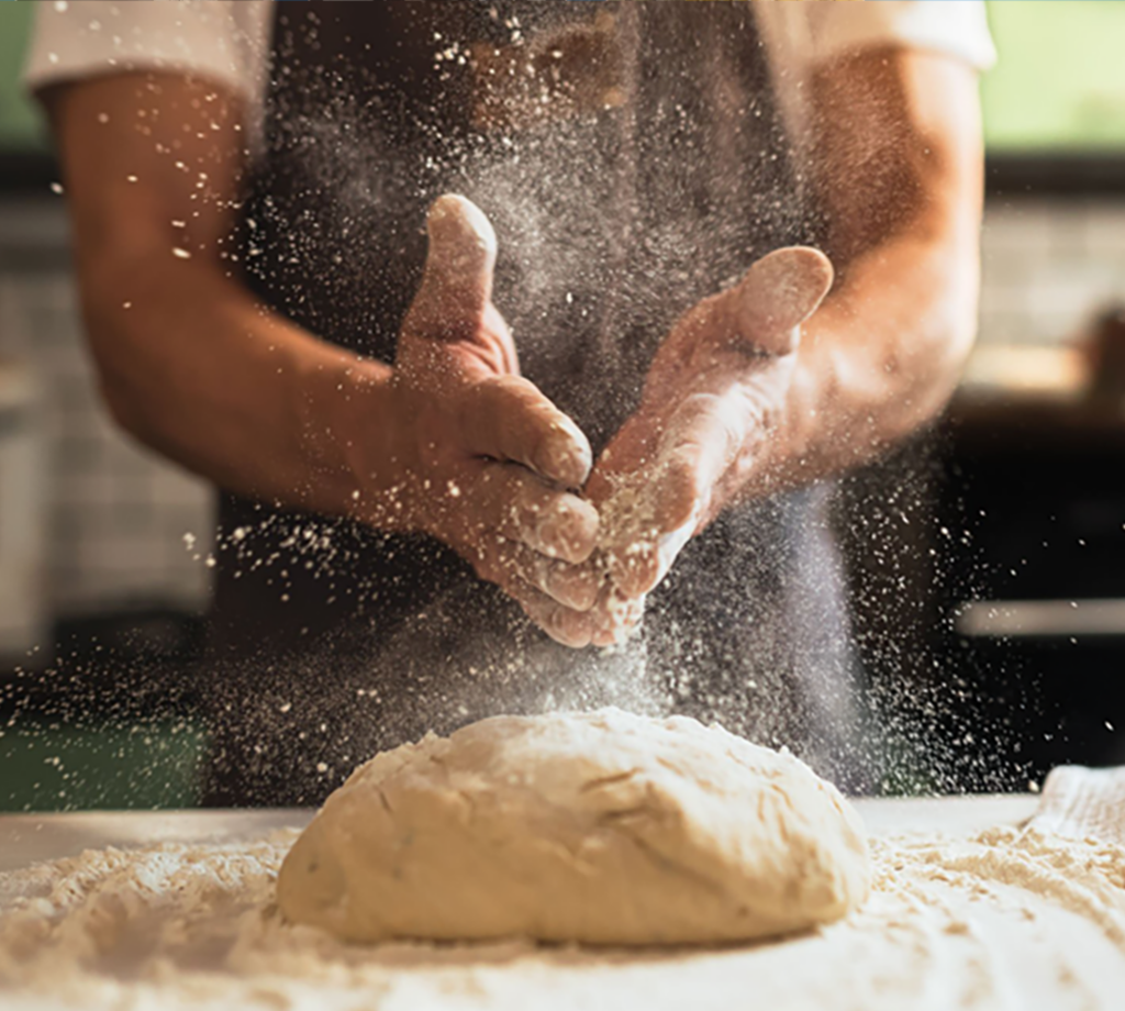 A person clapping their hands of flour over a pizza dough.