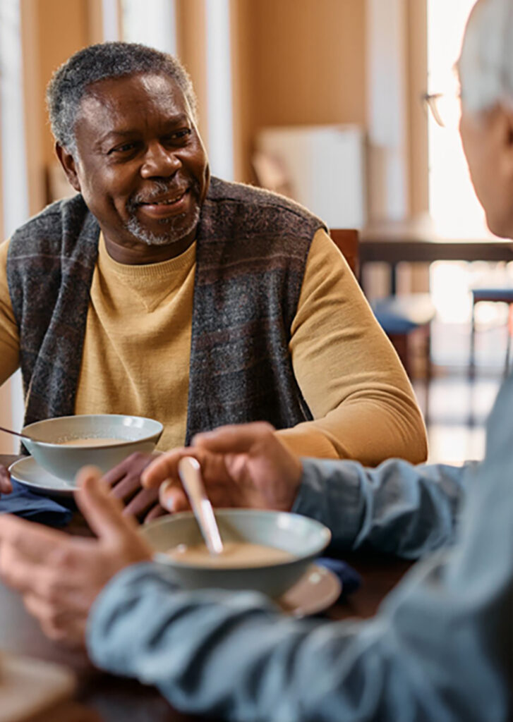 An African American sitting a table eating soup talking to another older man.