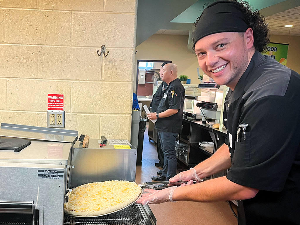 A NUT employee putting a cheese pizza in the oven.
